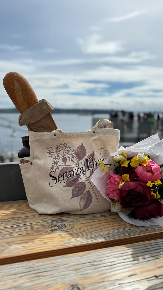 Beige tote bag with 'SennzaFinne' branding, bread loaf, and bouquet of flowers on a wooden surface with a waterfront background.