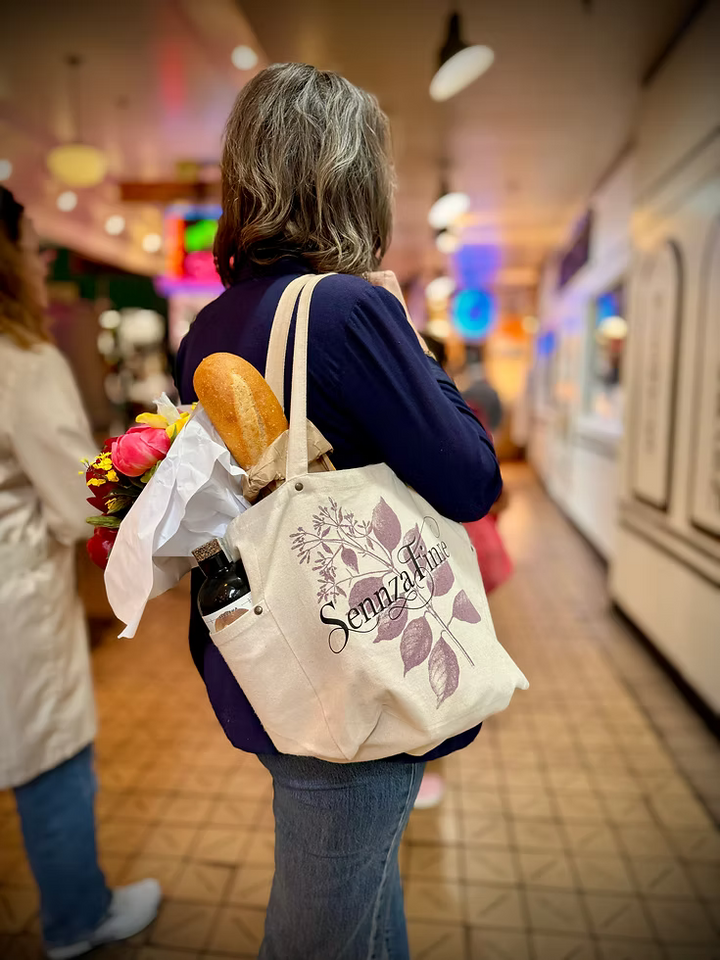Beige tote bag with 'SennzaFinne' branding, bread loaf, and bouquet of flowers on a wooden surface with a waterfront background.