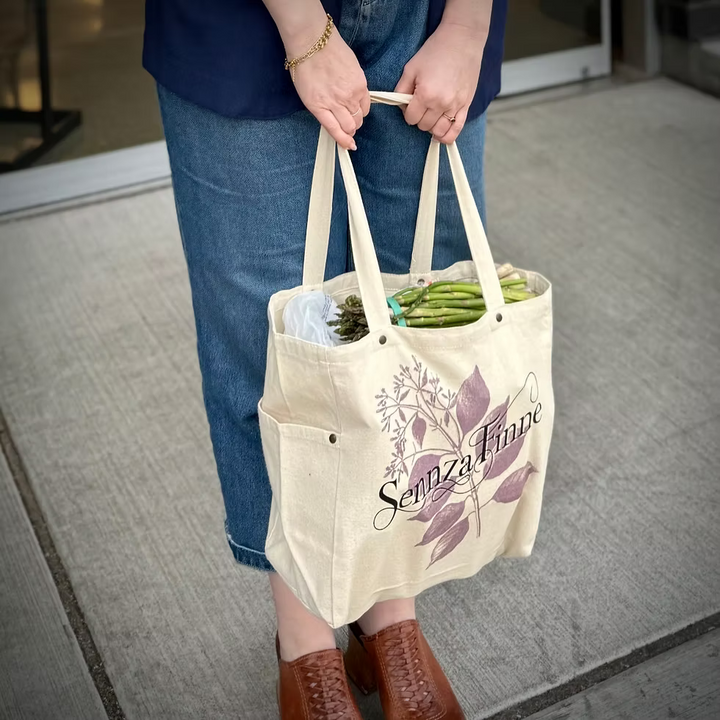 Beige tote bag with 'SennzaFinne' branding, bread loaf, and bouquet of flowers on a wooden surface with a waterfront background.